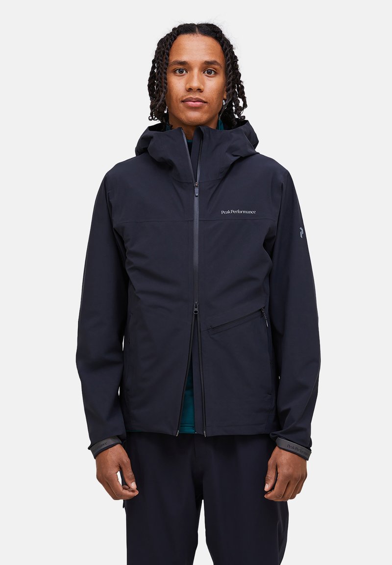 Giovane con trecce che indossa una giacca nera con cappuccio, aperta a metà, in piedi davanti a uno sfondo chiaro e uniforme.