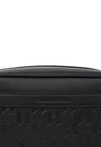 Svart lærveske med preget geometrisk mønster foran, glatt topp og glidelås-lukking. Merket med "KARL LAGERFELD."