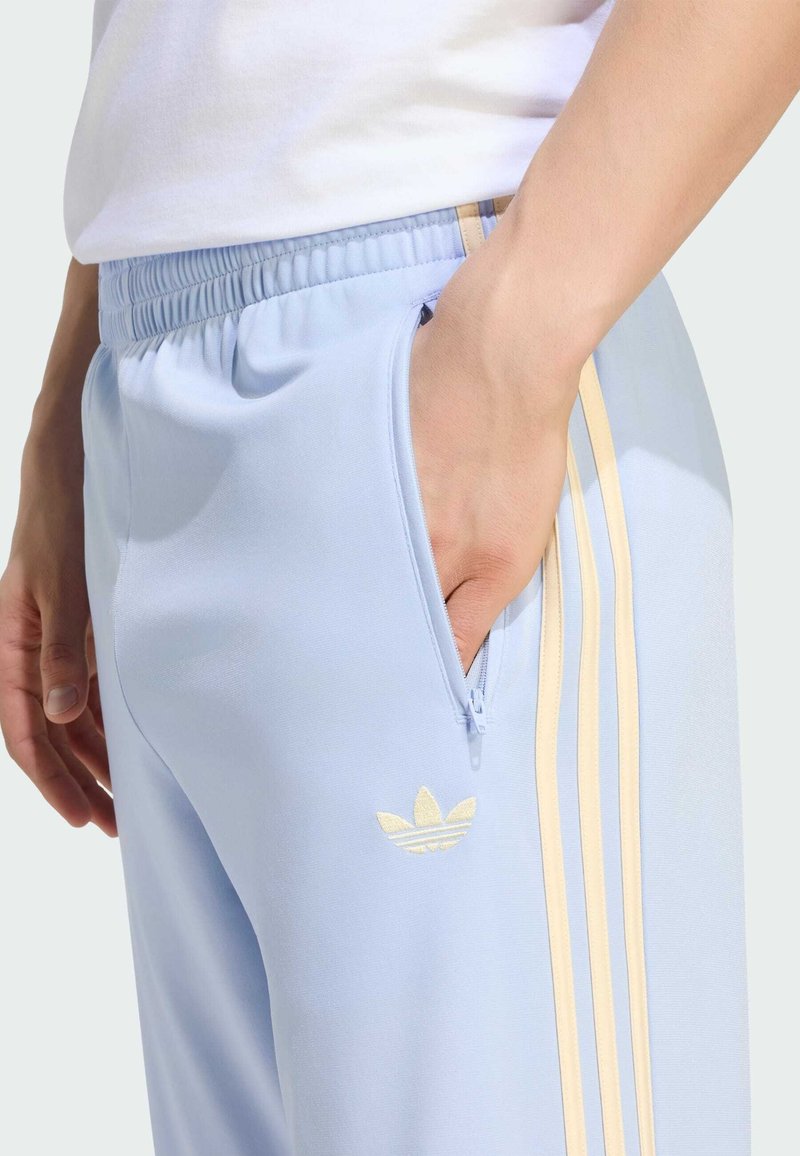 Person trägt hellblaue Adidas-Trainingshose mit beigen Streifen, elastischem Bund und Hand in seitlicher Reißverschlusstasche.