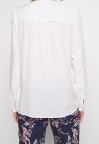 Blouse blanche à pois texturés, coupe ample, manches longues avec poignets boutonnés, et un bord en dentelle le long de la couture supérieure du dos.