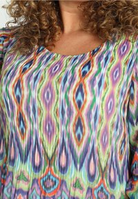 Blouse colorée présentant un motif multicolore avec des formes abstraites ondulées en violet, vert, bleu et orange. Tissu doux avec une légère texture.