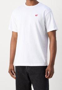 Weißes Baumwoll-T-Shirt mit kurzen Ärmeln, das auf der Vorderseite ein kleines rotes gesticktes Design aufweist. Kombiniert mit schwarzen Jeans.