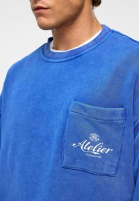 Blauwe katoenen sweatshirt met een ronde hals, lange mouwen en een zak met witte bedrukte tekst "Atelier Creative Department".