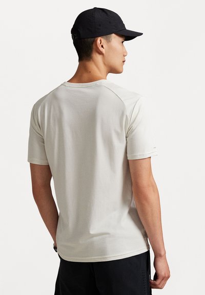 Νεαρός άνδρας με λευκό κοντομάνικο μπλουζάκι, μαύρο σορτς και μαύρη καπέλο του μπέιζμπολ, γυρισμένος προς τα πίσω σε λευκό φόντο.