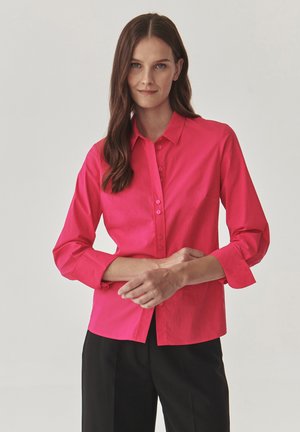 Camicetta rosa brillante a maniche lunghe con colletto, chiusura a bottoni e polsini arrotolati. Tessuto liscio e design aderente con orlo dritto.