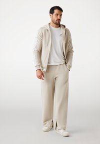 Beige Trainingsanzug mit einer Jacke mit Reißverschluss und Kapuze, die gestreifte Ärmel hat, kombiniert mit locker sitzenden Hosen und weißen Sneakern. Glattes Material.