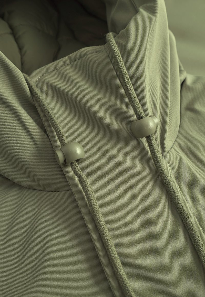 Chaqueta con capucha de color verde claro, que presenta una textura suave, cordones ajustables y un cierre con topes de plástico redondeados.