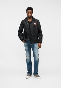 Νέος άνδρας που στέκεται όρθιος φορώντας μαύρο μπουφάν με το λογότυπο Diesel, ξεθωριασμένο μπλε τζιν και γκρι αθλητικά παπούτσια σε λευκό φόντο.