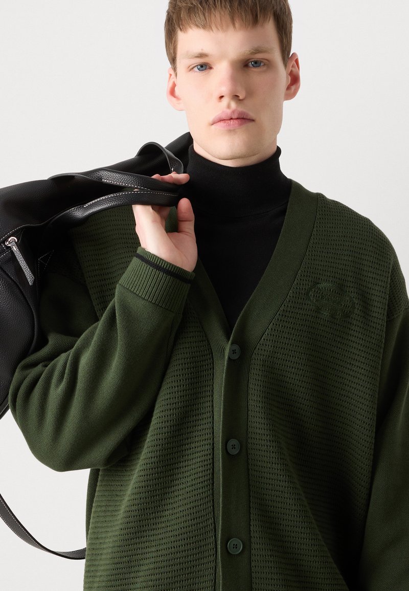 Zielony sweter z węzłami o teksturowanym wzorze, z trzema guzikami i ściągaczami u rękawów. Model trzyma czarną torbę na jednolitym tle.