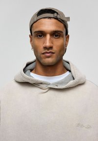 Beige hoodie met een getextureerde grijze voering, voorzien van een trekkoordkap en een geborduurd logo. Draagt met een wit T-shirt eronder.