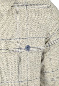 Beige geruite shirt gemaakt van textuurstof. Heeft een borstzak met een blauwe knop en een blauw ruitpatroon op een lichte achtergrond.