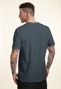 Tmavě šedé tričko s krátkým rukávem, hladká textura, kulatý výstřih a minimalistický design, zobrazené zezadu s viditelnými tetováními na paži.