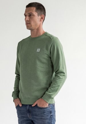 Mann trägt ein grünes strukturiertes Sweatshirt mit langen Ärmeln und kleinem Brustlogo und blaue Jeans, steht mit Händen in den Taschen.
