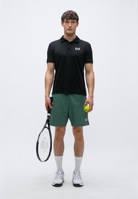 Juodas polo marškinėlis su logotipu, žali šortai, balti teniso bateliai, rankose geltonas teniso kamuoliukas ir raketė, stovintis prieš neutralią foną.