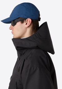 Svart vattentät jacka med hög krage och justerbar huva, i kombination med en blå keps och polariserade solglasögon, sedd från sidan.