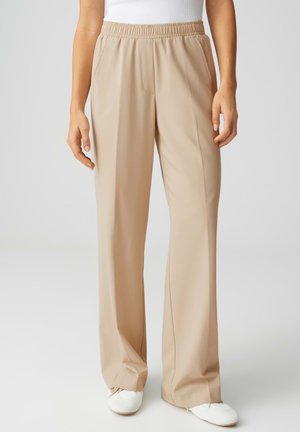 Beige weite Hosen mit elastischem Bund, getragen mit weißem Oberteil und weißen Schuhen, von der Taille bis zu den Füßen gezeigt.