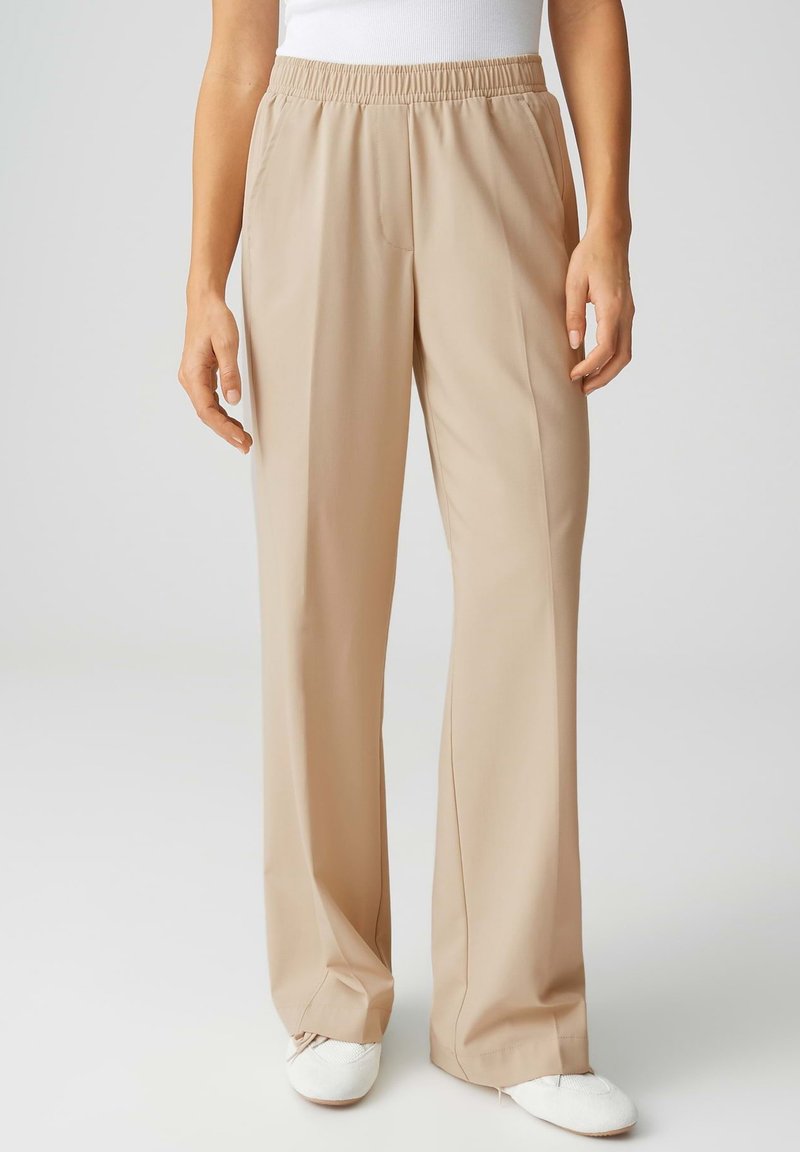 Beige wijde broek met elastische tailleband, gedragen met een wit topje en witte schoenen, weergegeven van taille tot voeten.