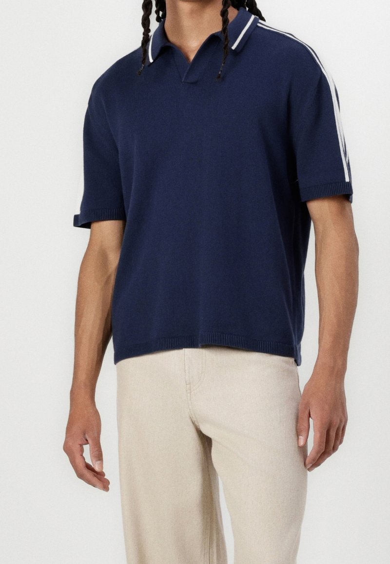 Homme portant un polo à manches courtes navy avec des rayures blanches sur les manches, associé à un pantalon beige clair, se tenant devant un fond uni.