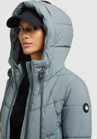 Svetlošedá prešívaná bunda s kapucňou, s predným zipsom, šnúrkovými detailmi a čiernou logom nášivkou na rukáve.