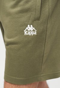 Λαδί πράσινα σορτς με πλαϊνή τσέπη και λευκό λογότυπο Kappa που απεικονίζει δύο καθιστές φιγούρες πλάτη με πλάτη κεντημένες στο αριστερό πόδι.