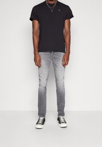 Camiseta de algodón negra de manga corta con un pequeño logo, combinada con jeans desgastados gris claro y zapatos negros con suelas blancas.