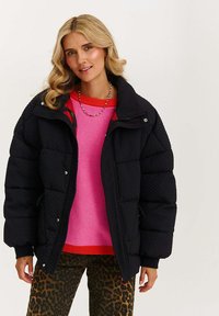 Žena s vlnitými blond vlasy, oblečená v černé prošívané bundě přes růžový svetr s červenými detaily a kalhoty s leopardím vzorem, pózuje před bílým pozadím.