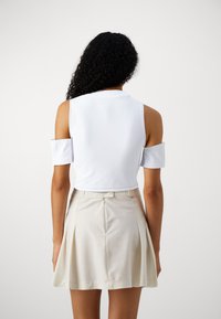 Λευκό κοντό μπλουζάκι με κοντά μανίκια, συνδυασμένο με μια μπεζ πλισέ φούστα. Η μπλούζα έχει κομψό σχέδιο με στρογγυλή λαιμόκοψη και ανοιχτές λεπτομέρειες στους ώμους.