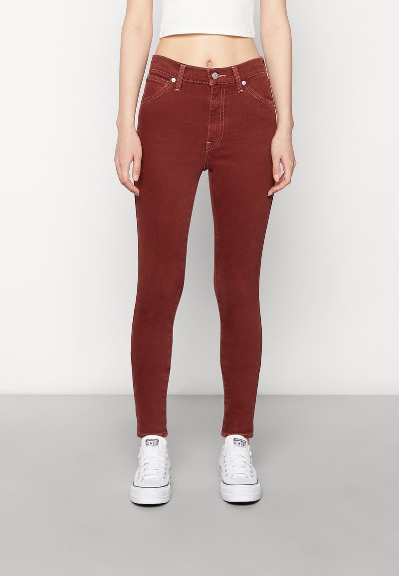 Femme portant un jean slim taille haute rouge foncé et des baskets blanches, debout devant un fond clair uni.