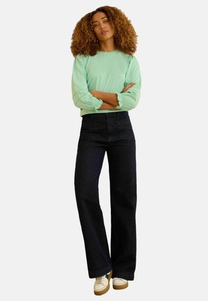 Femme aux cheveux bouclés portant un haut manches longues vert menthe, un pantalon large noir et des baskets blanches, debout les bras croisés.