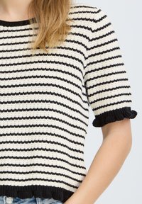 Femme portant un pull en maille à manches courtes avec des rayures horizontales noires et blanches et des bords noirs volantés aux manches et à l'ourlet.