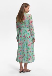 Genomskinlig klänning med långa ärmar i ljusgrön med ett färgglatt blommönster. Inkluderar en bälte i midjan och en fladdrig nederkant. Bärs med platta skor.