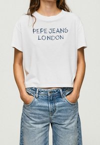 Camiseta corta blanca con "PEPE JEANS LONDON" impreso en azul marino. Combinada con jeans de denim de pierna ancha en azul claro. Diseño sencillo, estilo casual.