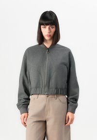 Harmaa lyhyt bomber-takki, jossa on sileä pinta, vetoketjusulkeminen ja ribbed-hihansuut. Etupuolella on pyöristettyjen sauma-akcenttien yksityiskohtia.