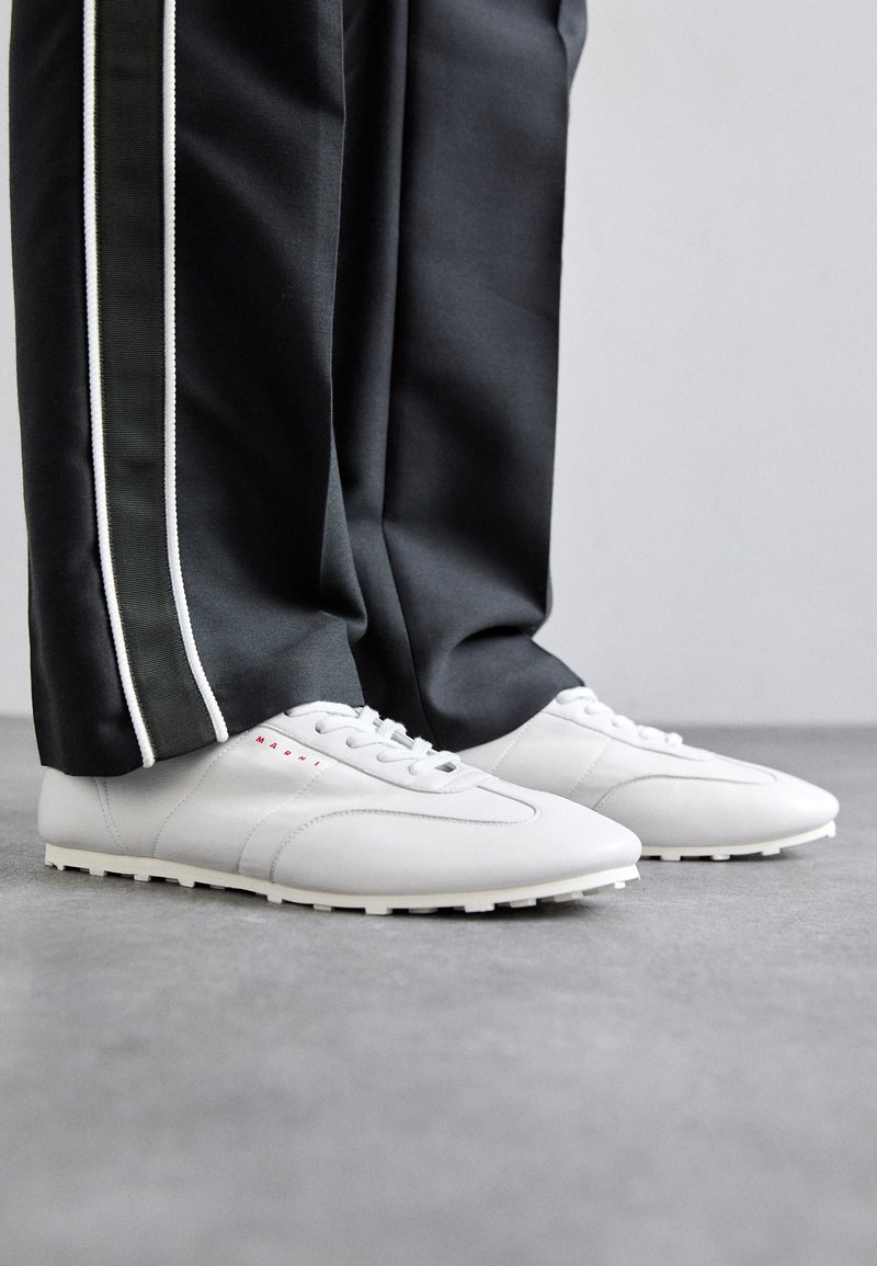 Witte veterschoenen op een grijze vloer, gedragen met zwarte broek met witte en zwarte zijkanten.