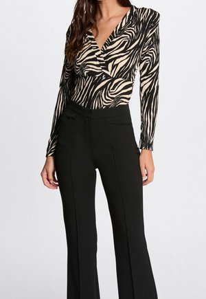 Kvinde iført en sort og beige zebra-mønstret V-hals bluse med lange ærmer, der er stoppet ned i højtaljede sorte bukser med syninger foran.