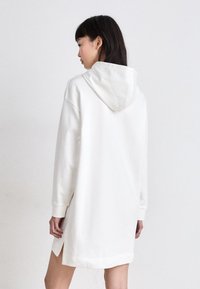 Bílá mikinová šatová šaty s dlouhými rukávy, bočními rozparky a jemnou texturou látky. Design je ležérní a oversized.