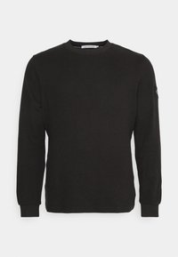 Sweatshirt noir à manches longues en mélange de coton. Texture côtes sur les poignets et le col. Présente une petite pièce logo sur la manche gauche.