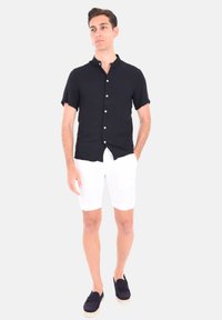 Camicia nera a maniche corte con bottoni e colletto rialzato, indossata con pantaloni corti bianchi e scarpe scivolate scure, in piedi su uno sfondo bianco.