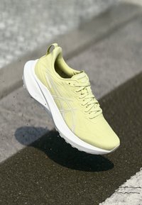 Baskets de course jaunes avec une tige texturée, des accents blancs, un bout rond et un col rembourré, affichées flottant au-dessus d'une surface pavée.