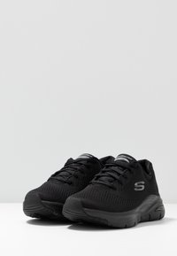 Svarta träningsskor med ovandel i mesh, rund tå, vadderad sula och texturerade detaljer. Har en logotyp på sidan och snörningsdesign.