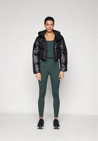 Črna krajšana puhasta jakna čez temno zeleno športno majico brez rokavov in ujemajoče se legice. Kompletu dodajo zaključek črni športni čevlji. Gladki materiali, sodoben kroj.