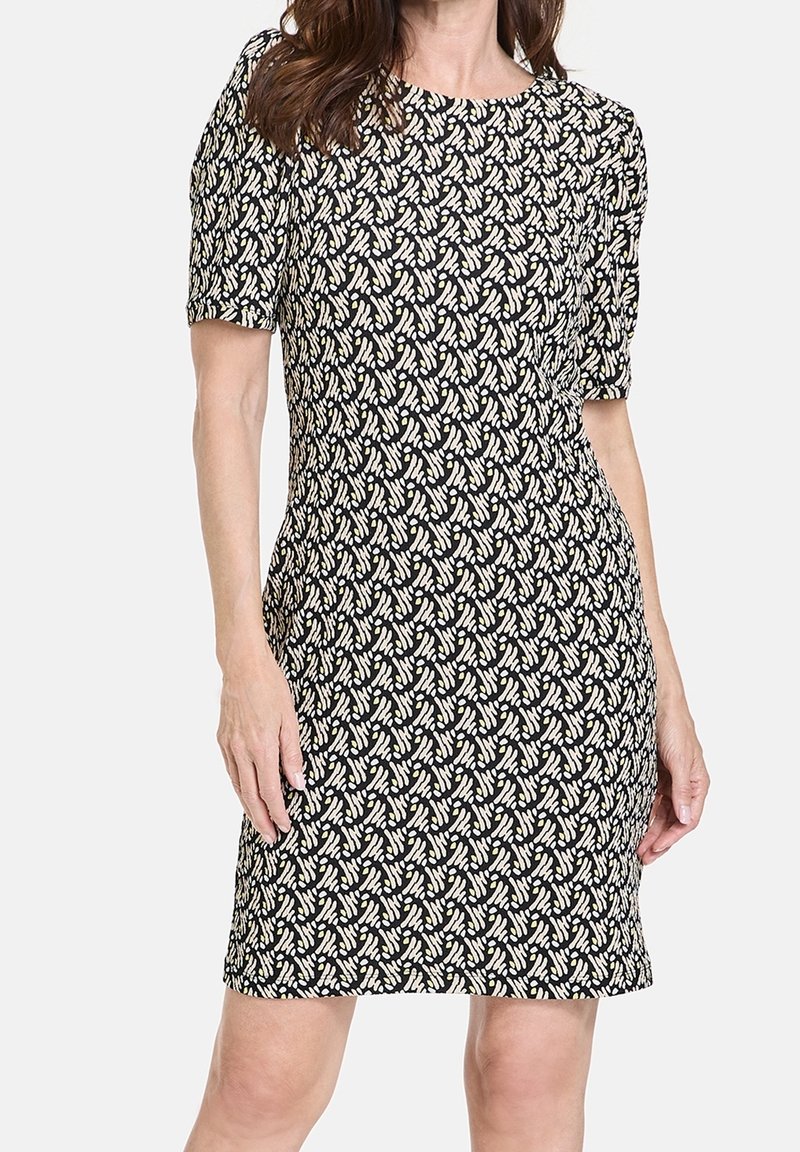 Femme portant une robe noire longueur genou avec des manches courtes et un motif abstrait blanc en forme de feuille, debout devant un fond uni.