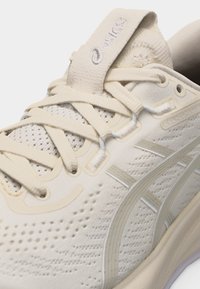 Κοντινό πλάνο μπεζ παπουτσιού για τρέξιμο Asics Gel-Cumulus με πλέγμα στο πάνω μέρος, κορδόνια και γλώσσα με λογότυπο.