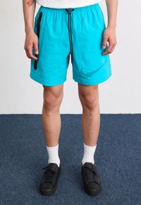 Turkise træningsshorts med sort lynlåslomme, snoreliv og prikket Nike-logo. Båret med hvide sokker og sorte sko.