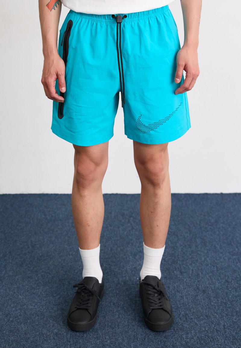 Turkise træningsshorts med sort lynlåslomme, snoreliv og prikket Nike-logo. Båret med hvide sokker og sorte sko.