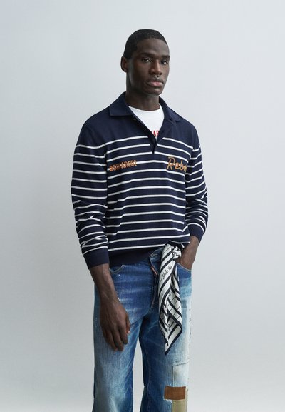 Homme portant un pull à rayures bleu marine et blanc, un jean bleu délavé avec une pièce, et une écharpe à motifs noir et blanc accrochée à une passants de ceinture.