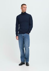 Hombre de pie con las manos en los bolsillos, vistiendo un suéter de cuello alto azul marino, jeans azules y zapatos negros, sobre un fondo claro liso.