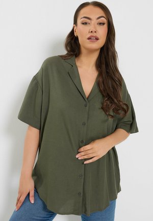 Donna con lunghi capelli castani che indossa una camicia verde oliva a maniche lunghe e jeans blu, posando contro una parete bianca e semplice.