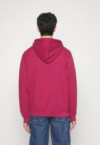 Felpa maroon con cappuccio e cordino, texture morbida, polsini e orlo a costine. Abbinata a jeans baggy blu. Vista da dietro.