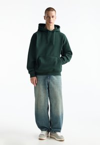 Grüner Kapuzenpullover mit einer Vordertasche, kombiniert mit lockeren hellblauen Jeans und grauen Sneakern, vor einem weißen Hintergrund.
